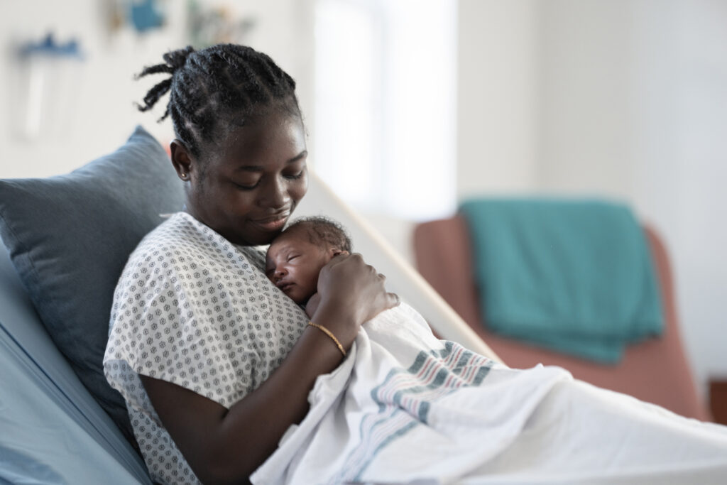 A Black mother gently holds her newborn close in a hospital setting, showcasing tender skin-to-skin bonding and peaceful, intimate family addition during the postpartum moment.